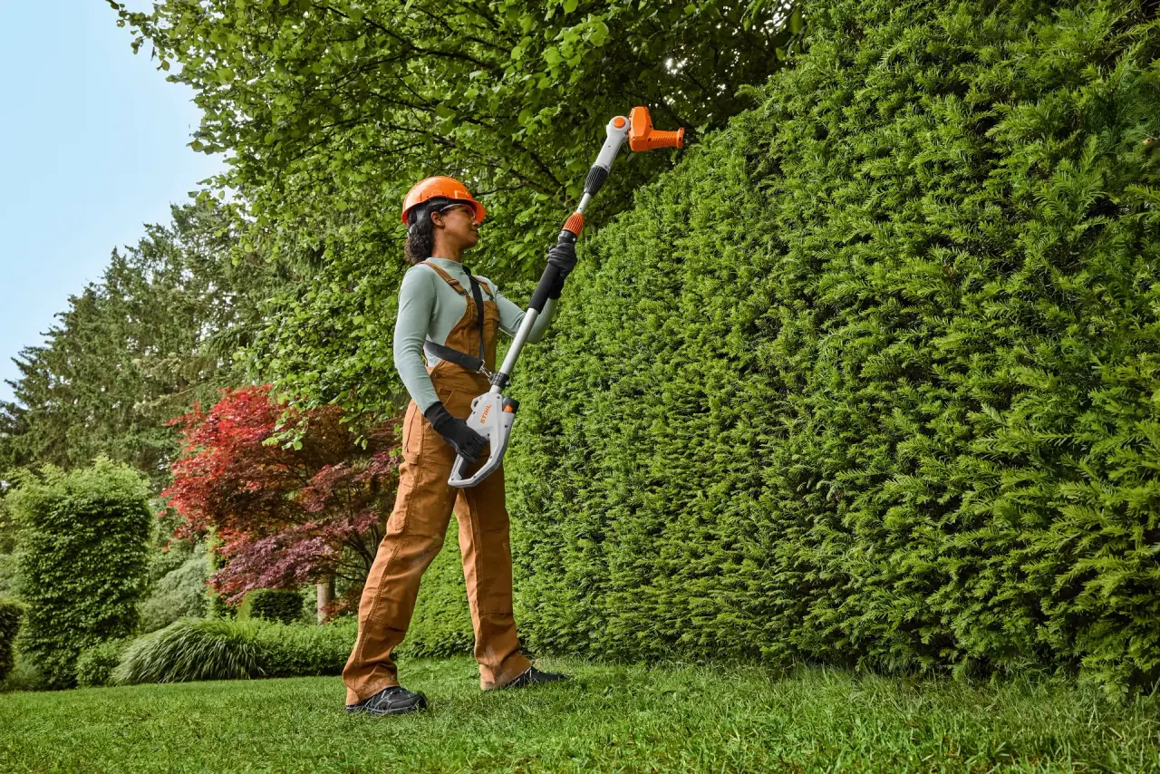 Gardener in protective gear trimming the top of a tall conifer hedge with a STIHL long-reach battery hedge trimmer